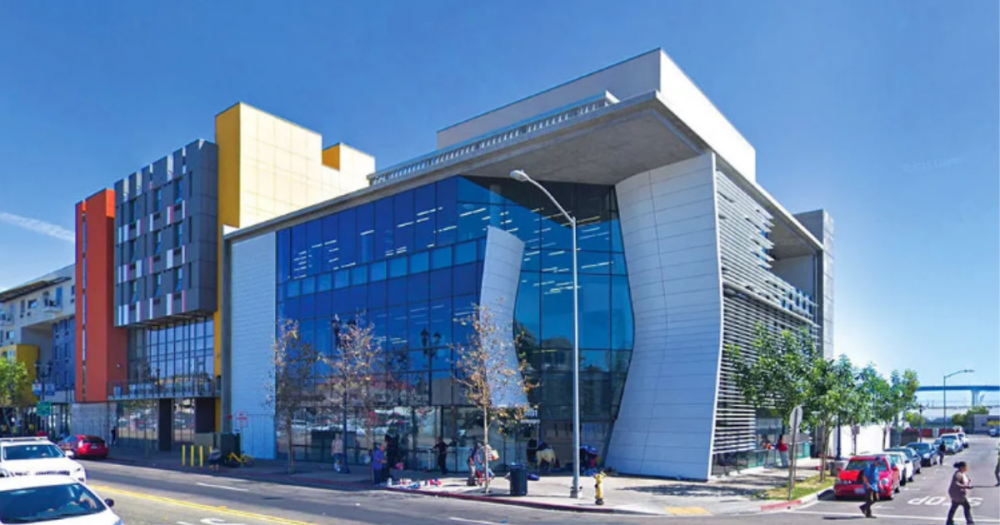 A glass building located at the corner of a street surrounded by people walking on the sidewalk and parked cars 