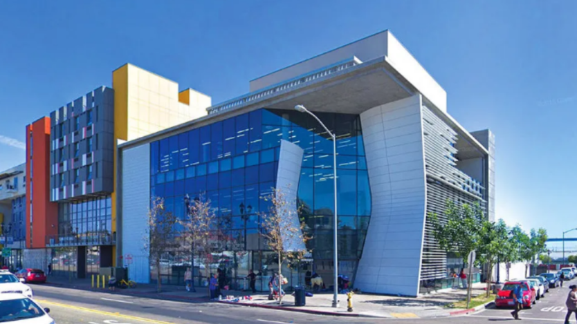 A glass building located at the corner of a street surrounded by people walking on the sidewalk and parked cars 